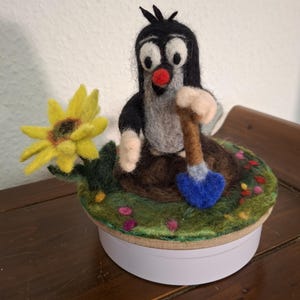 Pode incluir: Uma figura de feltro de um toupeira de desenho animado cavando com uma pá azul, colocada sobre uma base redonda com um gramado verde feltrado e um girassol amarelo. A toupeira é preta, cinza e branca com um nariz vermelho.