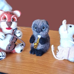 Puede incluir: Un conjunto caprichoso de animales músicos de fieltro. Un ratón blanco toca la guitarra, un zorro rojo y blanco toca el violín, una nutria gris toca el saxofón y un ratón blanco toca el piano. Cada animal lleva una falda y está sobre una superficie de madera.