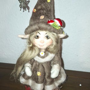 Könnte beinhalten: Eine handgefertigte Zwergenpuppe aus Filz mit einem skurrilen Design. Der Zwerg trägt einen braunen Hut mit Blättern und Pilzen, ein braunes Kleid und hat lange blonde Haare. Ein Schmetterlingsanhänger hängt am Hut.