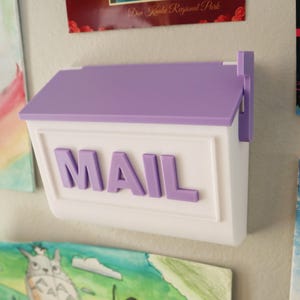 May include: A white mailbox with a purple roof and the word "MAIL" in purple letters. The mailbox is mounted on a wall with artwork in the background.