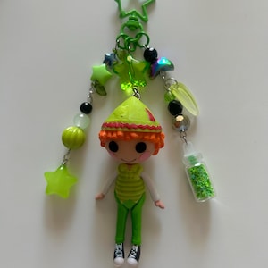 May include: A keyring featuring a small doll with orange hair wearing a green outfit and hat. The doll is attached to a green star-shaped clasp with various green, black, and iridescent beads, star charms, and a small clear bottle filled with green glitter.