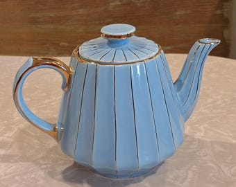 Può includere: Una teiera in ceramica azzurra con coperchio, manico e beccuccio. La teiera presenta strisce verticali dorate e finiture dorate sul manico, sul beccuccio e sul coperchio. La teiera è su una superficie bianca.