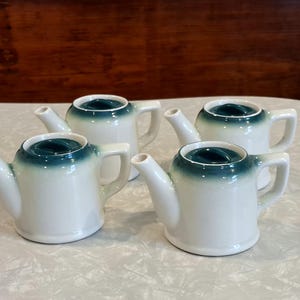 May include: Four small white ceramic teapots with green lids. The teapots have a rounded body and a small spout.