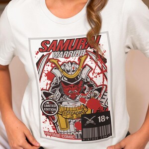 May include: White t-shirt featuring a vintage comic book style graphic. The design includes a samurai warrior with a red mask and helmet, set against a backdrop of red splatters. Text includes "SAMURAI WARRIOR", "LIMITED EDITION", and "18+".
