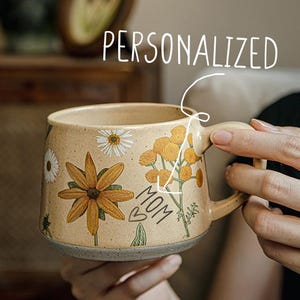 Può includere: Una tazza in ceramica beige con un motivo floreale e la parola "MOM" scritta su di essa. La tazza ha un manico ed è personalizzata.