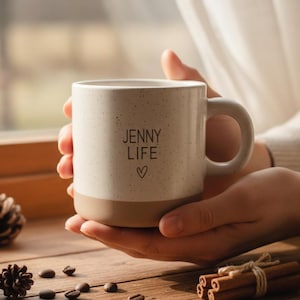 May include: A speckled ceramic mug with the words "JENNY LIFE" and a heart symbol. The mug is held by a person. The mug has a tan base and a white upper portion. Coffee beans, pine cones, and cinnamon sticks are on a wooden surface.