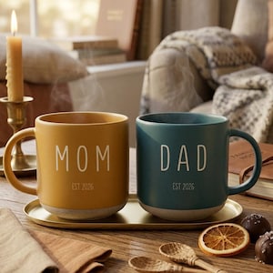 May include: Two ceramic mugs, one mustard yellow and one teal, sit on a gold tray. The yellow mug reads "MOM EST 2026" and the teal mug reads "DAD EST 2026". The mugs are on a wooden table with spoons, a candle, and a dried orange slice.