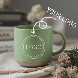 Puede incluir: Una taza de cerámica verde con una base marrón claro. La taza tiene un asa y un círculo blanco con la palabra "LOGO" en su interior. El texto "YOUR LOGO" está encima de la taza. La taza está sobre una tela blanca con flores secas y un palo de madera.
