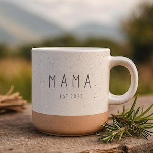 Può includere: Una tazza in ceramica bianca macchiata con la scritta "MAMA" e "EST. 2025" stampata in grigio. La base della tazza è color beige. La tazza ha un manico e si trova su una superficie di legno.