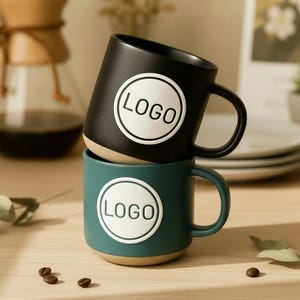 Puede incluir: Dos tazas de cerámica, una negra y otra verde azulado, apiladas una encima de la otra. Cada taza tiene un círculo blanco con la palabra "LOGO". Las tazas tienen una base beige y un asa curvada. Granos de café y una cafetera de cristal en el fondo.
