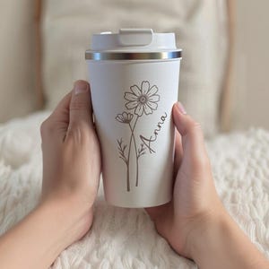 Op de afbeelding: Een witte reisbeker met een zilveren rand en een wit deksel, voorzien van een donkerbruine lijntekening van twee wilde bloemen en de naam Anna in cursief schrift op de voorzijde, vastgehouden in twee handen tegen een textuurrijke achtergrond.
