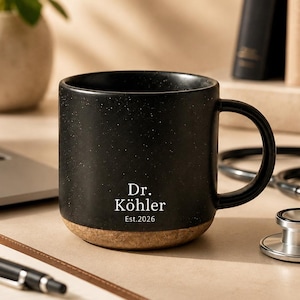 Könnte beinhalten: Ein schwarzer Keramikbecher mit gesprenkelter Oberfläche und einem Boden aus Naturkork. Auf der Vorderseite befindet sich ein weißer Aufdruck mit dem Text Dr. Köhler und Est. 2026. Der Becher hat einen geschwungenen Henkel und eine matte Textur.