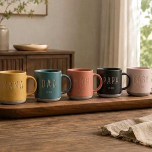 Könnte beinhalten: Fünf Keramiktassen in Gelb, Petrol, Koralle, Schwarz und Hellrosa mit eingeprägtem Text wie MAMA, DAD, MOM, PAPA und BABY sowie der Jahreszahl EST 2026 darunter. Jede Tasse hat einen grauen Boden und einen passenden Henkel.