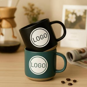 Puede incluir: Dos tazas de cerámica apiladas. La taza superior es negra y la inferior es verde azulado. Cada taza tiene un círculo blanco con la palabra "LOGO" impresa en negro. Las tazas tienen una base beige y un asa negra.