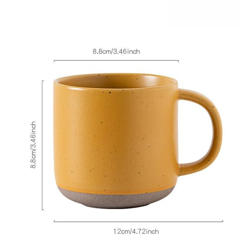 May include: Mustard yellow ceramic mug with a gray base. The mug is 4.72 inches wide and 3.46 inches tall. The mug has a large handle and a speckled texture. The mug is suitable for coffee, tea, or other beverages.