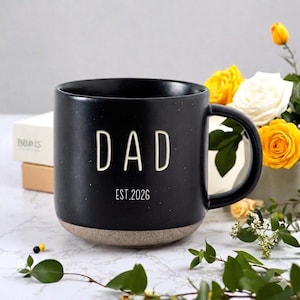 May include: Black ceramic mug with the word "DAD" in white block letters and "EST.2026" printed below. The mug has a speckled finish and a tan base. A bouquet of yellow and white roses is in the background.