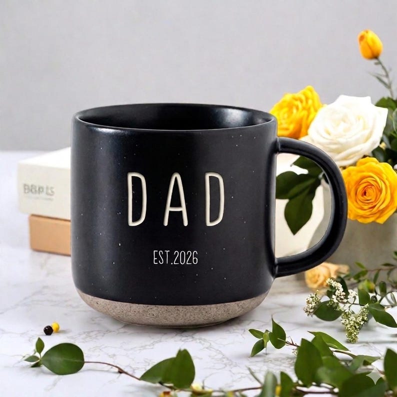 May include: Black ceramic mug with the word "DAD" in white block letters and "EST.2026" printed below. The mug has a speckled finish and a tan base. A bouquet of yellow and white roses is in the background.