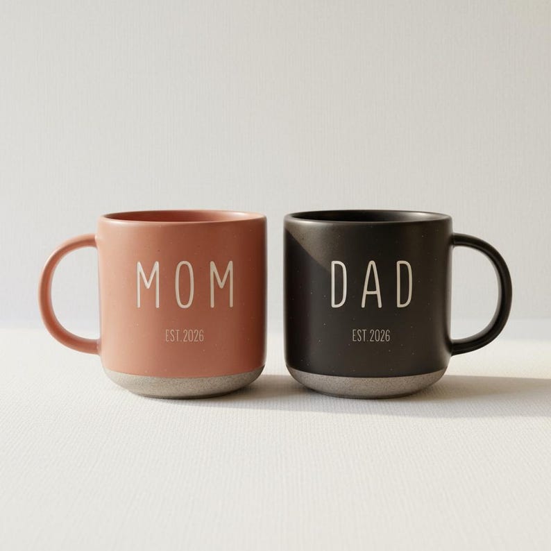 May include: Two ceramic mugs, one coral and one black, with the words "MOM" and "DAD" printed in white. Each mug has a gray base and the text "EST. 2026" below the main word. The mugs have a simple, modern design.