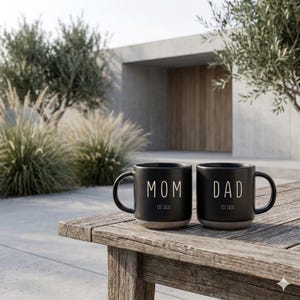 May include: Two black ceramic mugs with the words "MOM" and "DAD" in white lettering. Each mug has "EST 2026" printed below the text. The mugs are sitting on a weathered wooden table outdoors.