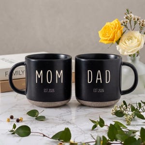May include: Two black ceramic mugs with the words "MOM" and "DAD" in white lettering. Each mug has "EST. 2026" printed below the main text. The bottom of the mugs are a light brown color.