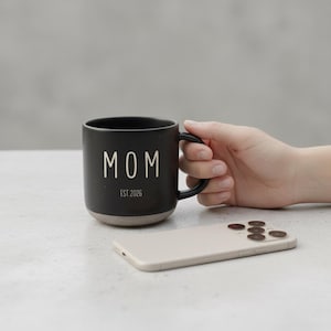 May include: A black ceramic mug with the word "MOM" in white lettering and "EST 2026" printed below. The mug has a black handle and a beige base. A beige smartphone is next to the mug.
