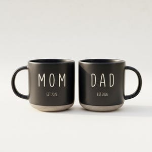 May include: Two black ceramic mugs with white text that reads "MOM" and "DAD". The mugs have a speckled finish and a gray base. The text "EST 2026" is below each word.