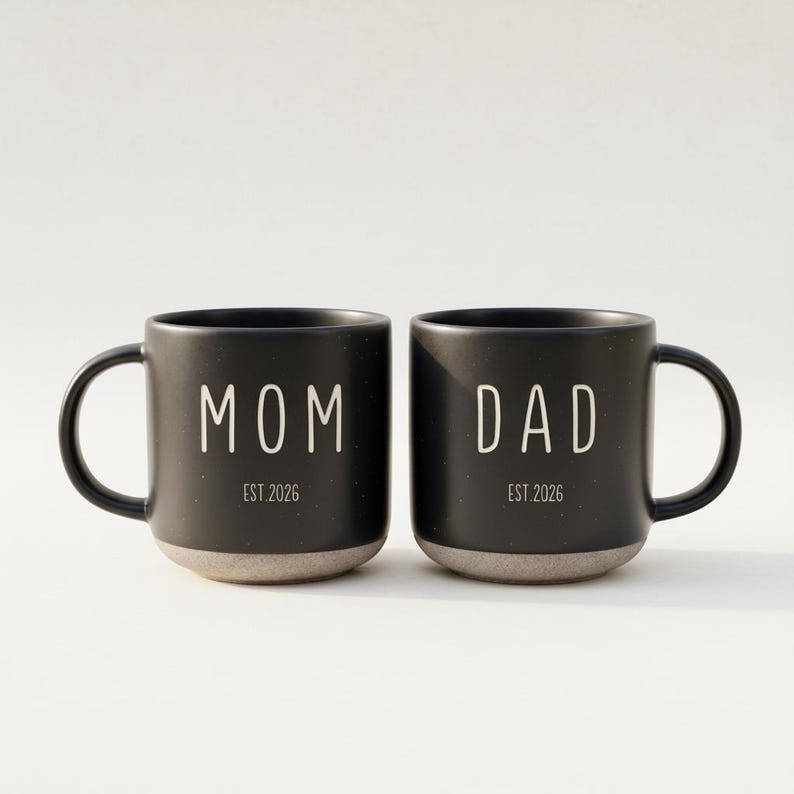 May include: Two black ceramic mugs with white text that reads "MOM" and "DAD". The mugs have a speckled finish and a gray base. The text "EST 2026" is below each word.
