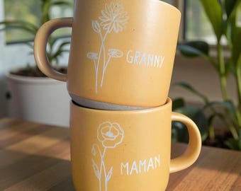 May include: Two yellow ceramic mugs stacked on top of each other. The top mug has the word "GRANNY" and a floral design in white. The bottom mug has the word "MAMAN" and a floral design in white. Both mugs have a handle.
