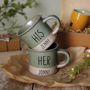 Peut inclure: Deux tasses en céramique vertes avec des accents blancs. La tasse du haut dit "HIS" avec le nom "JENNY" en dessous. La tasse du bas dit "HER" avec le nom "JONNY" en dessous.