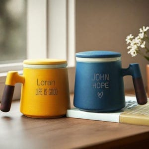 Op de afbeelding: Twee keramische mokken met deksels en houten handvatten. De gele mok zegt "Loran Life is Good" en de blauwe mok zegt "John Hope" met een hartje.