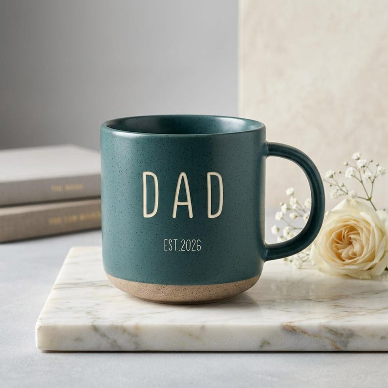 May include: A teal ceramic mug with the word "DAD" in white lettering. The mug has a speckled finish and a tan base. Below the word "DAD" is the text "EST. 2026". The mug is on a marble surface.