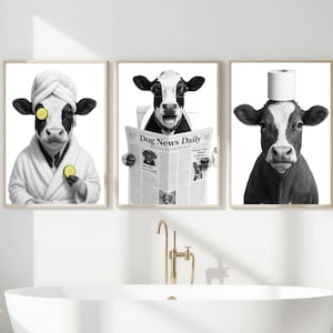 May include: Set of three framed black and white cow art prints. One cow wears a spa robe and cucumber slices on its eyes. Another cow reads a newspaper titled "Dog News Daily." The third cow has a roll of toilet paper on its head.