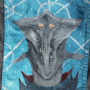 May include: A denim jacket with a hand-painted design of a gray alien with orange eyes on the back pocket. The alien is surrounded by white lines on a blue background.