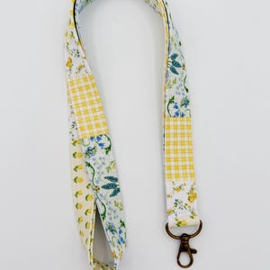 May include: A floral patterned lanyard with a bronze-colored clasp. The lanyard features a patchwork design with yellow gingham, white and yellow floral, and blue floral patterns. The lanyard is made of fabric.