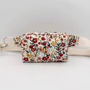 May include: A floral print fanny pack with a cream-colored adjustable strap. The bag features a white background with a pattern of red, yellow, and pink flowers and green leaves. The zipper is silver.