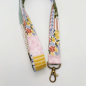 May include: A patchwork fabric lanyard with a bronze-colored swivel clasp. The lanyard features a variety of floral and patterned fabrics in shades of pink, yellow, blue, and green. The design is a mix of floral prints and stripes.