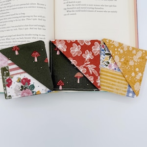 May include: Three handmade fabric corner bookmarks with different patterns are displayed open on a book. The patterns include mushrooms, floral designs, and stripes. The bookmarks are in shades of green, red, and yellow.