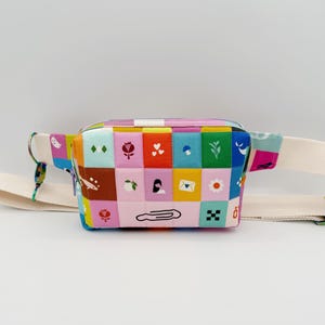 May include: A colorful fanny pack with a patchwork design featuring squares of various colors and patterns. The bag has a cream-colored adjustable strap and a rainbow-colored zipper pull. The design includes floral and geometric motifs.