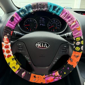 May include: A fabric steering wheel cover featuring a patchwork design with various Halloween-themed patterns including orange pumpkins, black cats, purple bats, yellow spiders, and owls on a dark background, installed on a black Kia steering wheel.
