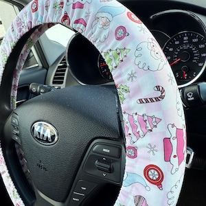 May include: A pink and white Christmas-themed steering wheel cover. The cover features a pattern of Santa Claus, Christmas trees, ornaments, and candy canes. The cover is on a black steering wheel in a Kia.