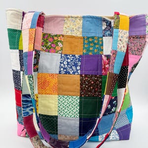 May include: A patchwork tote bag with a variety of colorful fabric squares. The bag features a mix of floral, solid, and patterned fabrics in shades of blue, pink, green, yellow, and purple. The straps are also made of patchwork fabric.