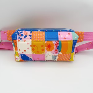 May include: A quilted fanny pack featuring a patchwork design of various colorful fabric squares. The patterns include a blue bird, a stylized face, floral motifs, and geometric shapes in shades of pink, yellow, orange, and blue. It has a blue zipper.