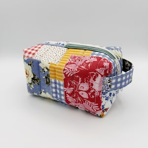 May include: A rectangular quilted pouch featuring a patchwork design of various fabrics, including floral patterns in blue, red, and white, yellow stripes, and blue gingham checks. The pouch has a zipper closure and a matching fabric side handle.