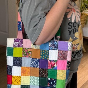 May include: A patchwork tote bag with a variety of colorful fabric squares. The bag has a rectangular shape with a matching strap. The fabrics include floral, solid, and patterned designs in shades of pink, blue, green, and purple.