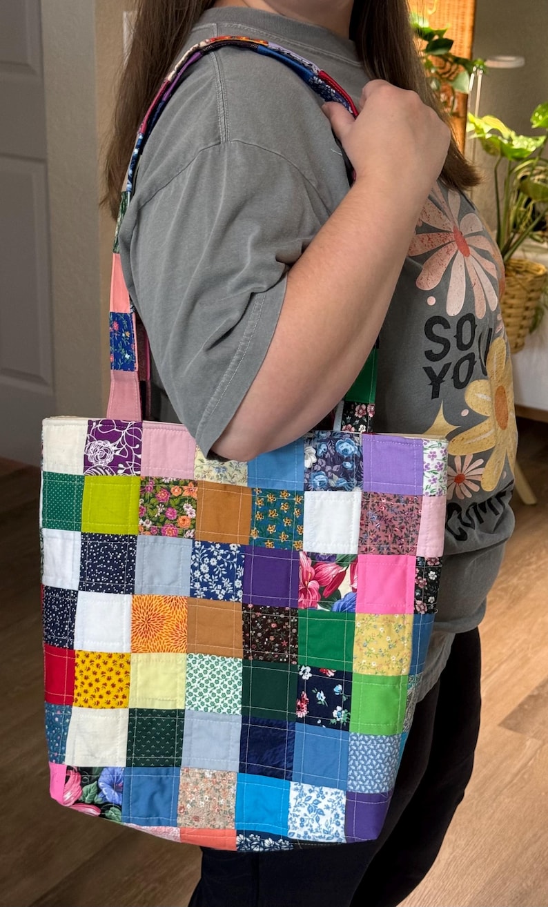 May include: A patchwork tote bag with a variety of colorful fabric squares. The bag has a rectangular shape with a matching strap. The fabrics include floral, solid, and patterned designs in shades of pink, blue, green, and purple.