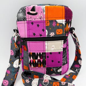 May include: A quilted crossbody bag featuring a patchwork design with Halloween-themed fabric patterns. The bag displays motifs of ghosts, pumpkins, witch hats, bats, and spiderwebs in shades of purple, orange, black, gray, and white with a black zipper.