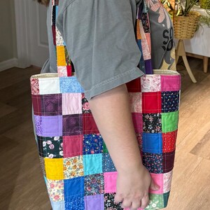 May include: A colorful patchwork tote bag with a variety of fabric squares in shades of pink, blue, yellow, and green. The bag has long straps and a rectangular shape, perfect for carrying items. The bag is handmade.