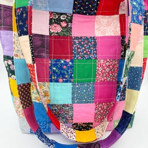 May include: A patchwork tote bag with a variety of colorful fabric squares. The bag features a mix of floral, solid, and patterned fabrics in shades of pink, blue, green, and purple. The handles are also made of patchwork fabric.