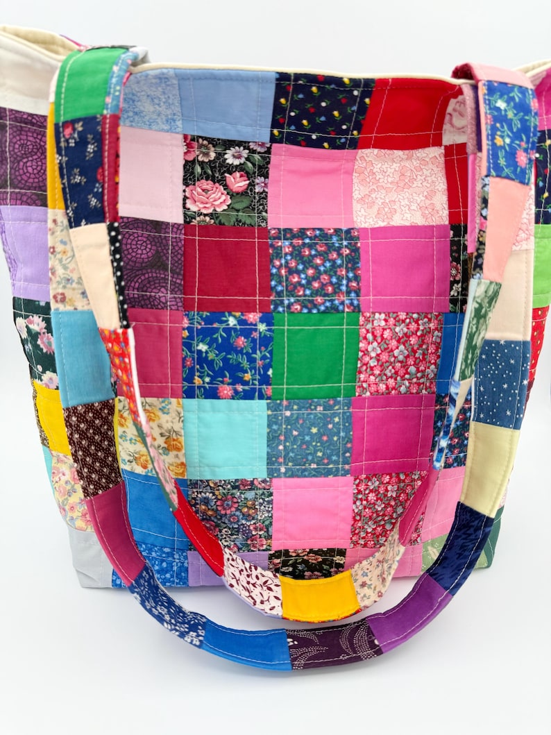 May include: A patchwork tote bag with a variety of colorful fabric squares. The bag features a mix of floral, solid, and patterned fabrics in shades of pink, blue, green, and purple. The handles are also made of patchwork fabric.