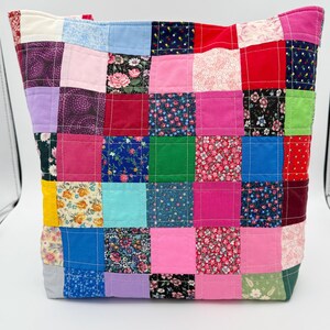 May include: A patchwork tote bag with a variety of colorful fabric squares. The squares feature floral, solid, and patterned designs in shades of pink, blue, red, green, and purple. The bag has a rectangular shape.